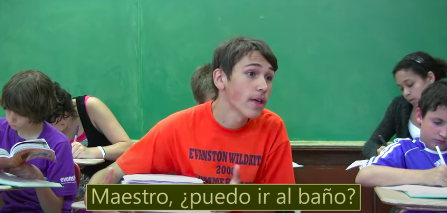 A student in a classroom sings to his teacher "Can I go to the bathroom" in spanish.