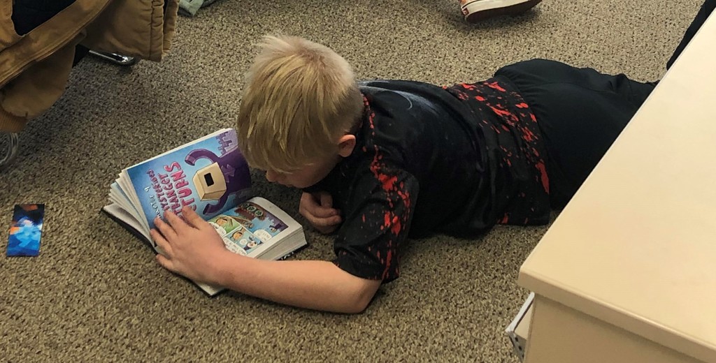 Boy lays on his tummy on the floor reading a book