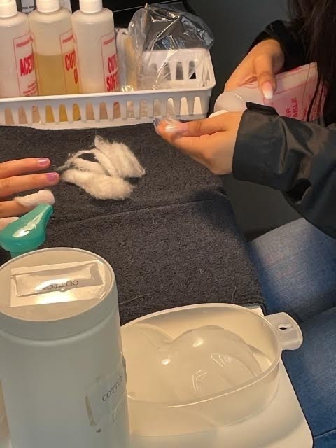 2 students, one with cotton on her finger nail while the other preps a cotton ball to place on her next finger. 