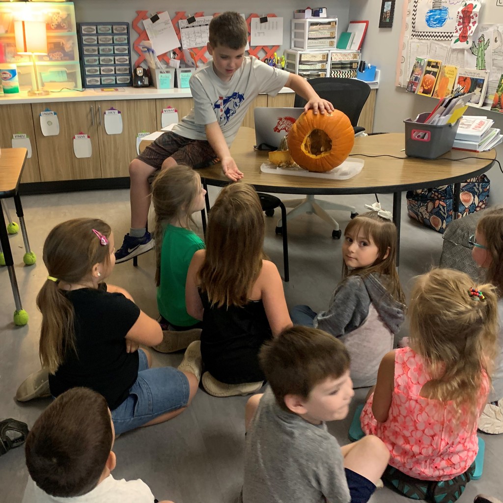 boy sitting on table pulling seeds out of  pumpkin with  whole in the side while other kids watch 