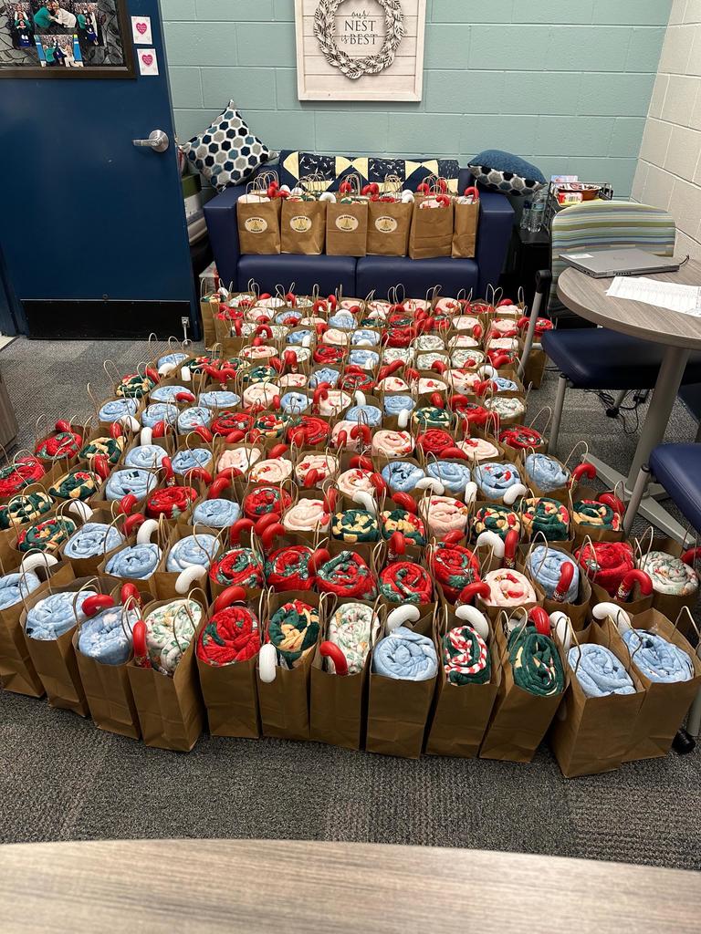Rows of gift bags filled with rolled blankets and small items arranged on the floor and a couch for a school event or donation project.