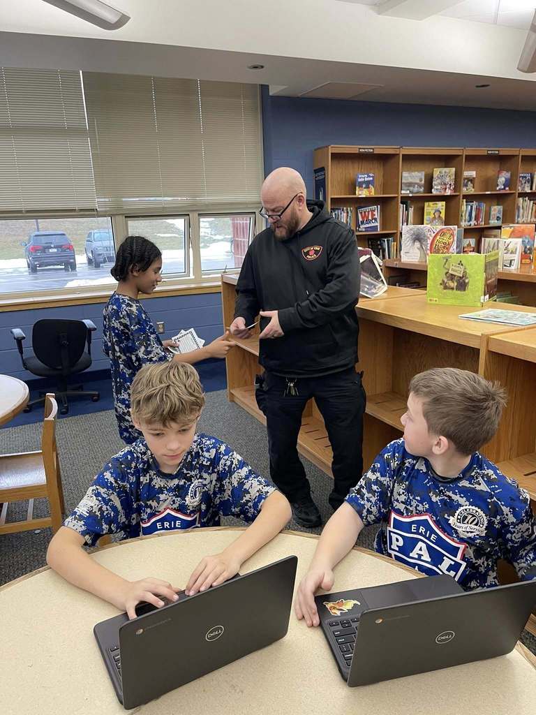 Students in a library and a student and police officer are handing each other something. 