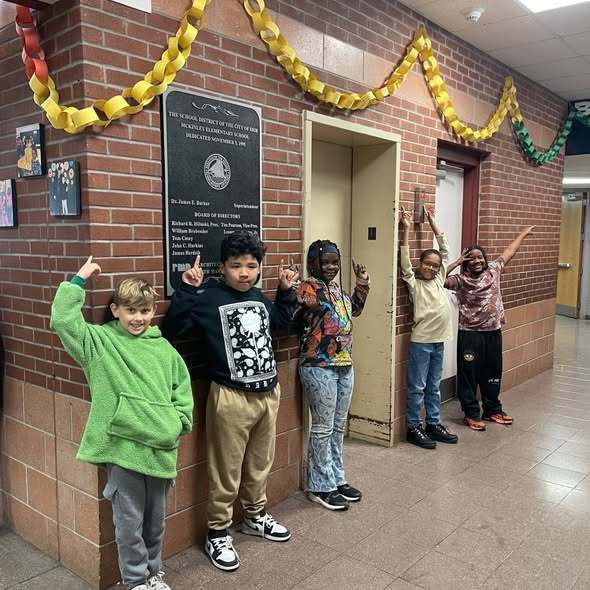 Kids standing in a hall and pointing up at a yellow paper chain. 