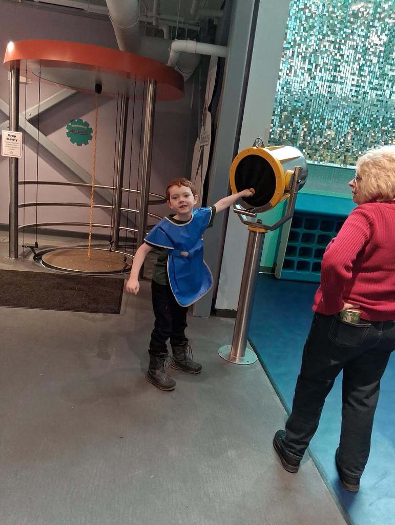 Young boy in a blue  apron holding his hand out to an object. 