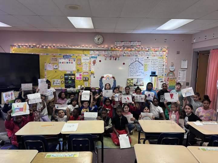A group of students in a classroom sitting and holding up drawings.