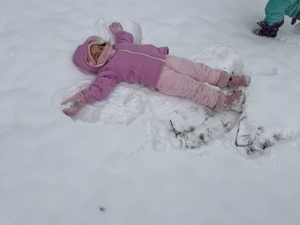 Young child in a pink winter outfit making a snow angel.