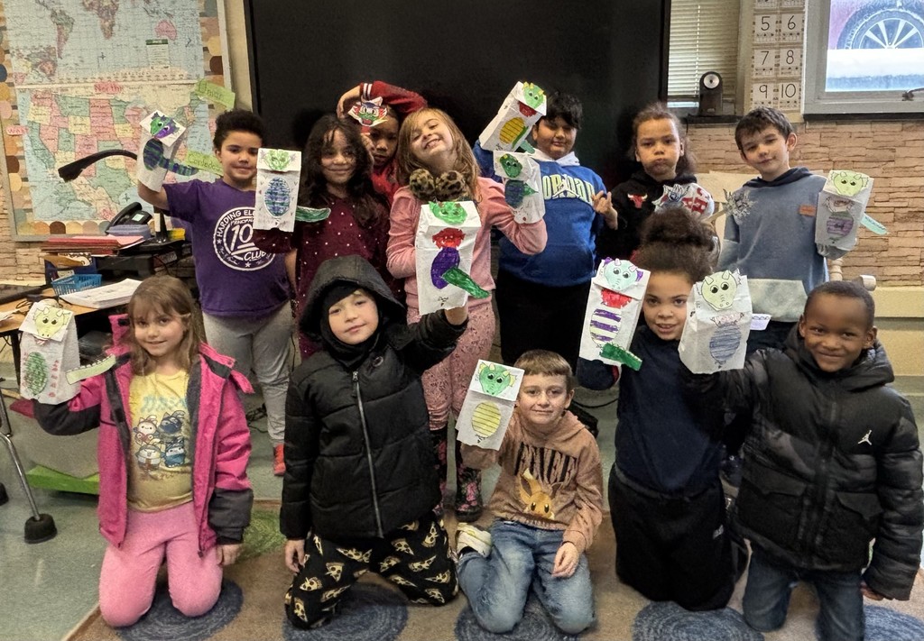 Students in a classroom wearing jackets and holding up dragon puppets they made. 