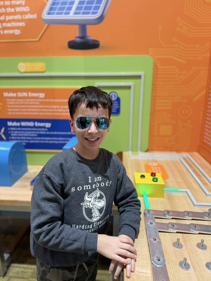 Young boy in a gray sweater smiling and wearing sunglasses. 