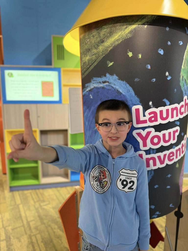 Young boy in a blue hoodie holding his pointer finger up and wearing glasses. 