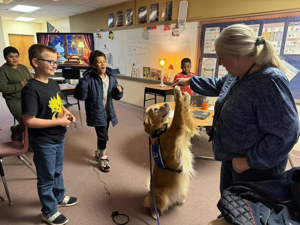 Kids smiling in a classroom, while a golden retriever dog does a trick. 