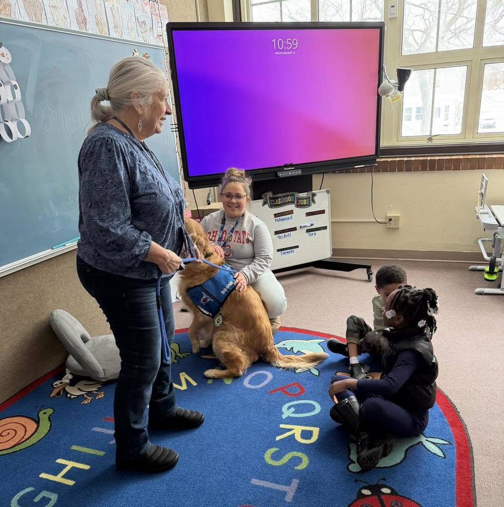 Female adult standing while kids are sitting on the floor in a classroom. 