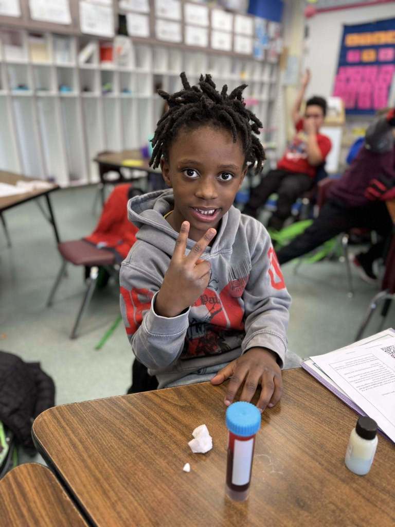 Young boy in a gray hoodie smiling and holding two fingers up. 