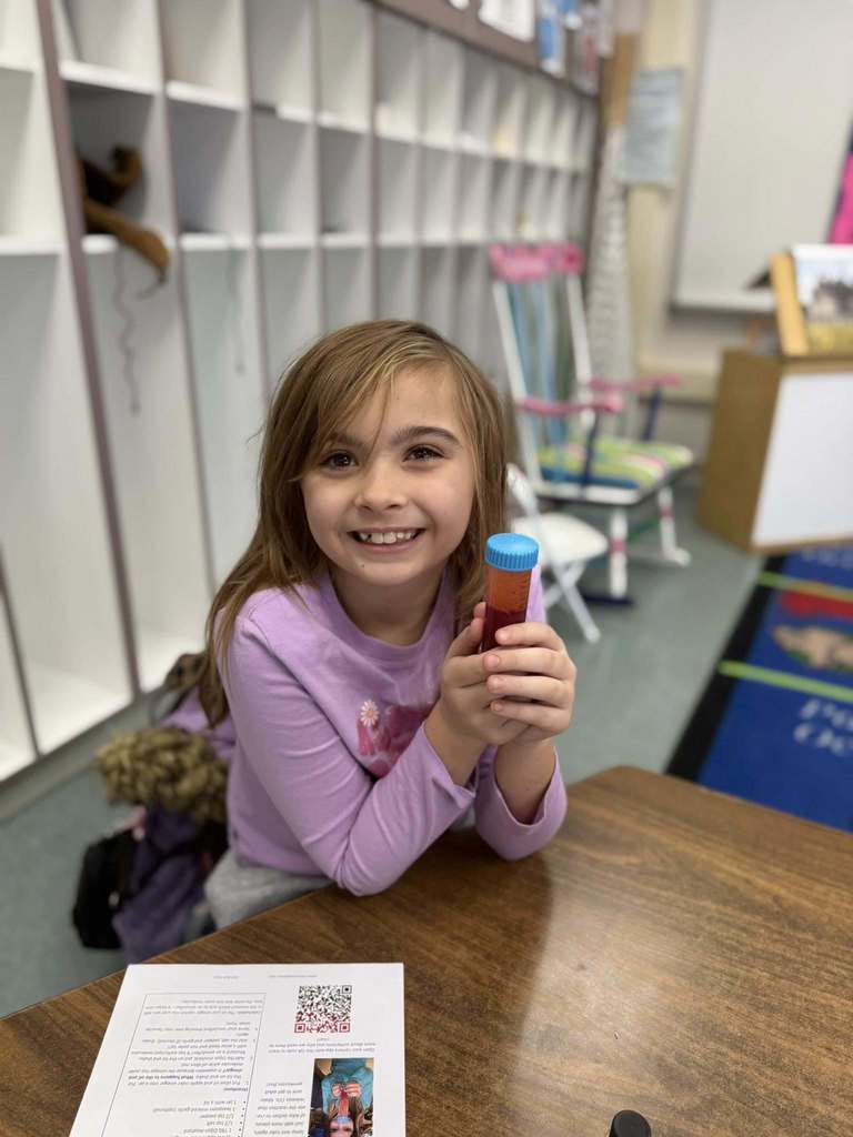 Young girl in a purple shirt smiling and holding a tube of red liquid. 