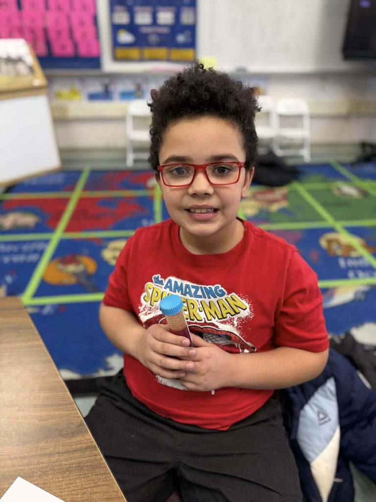 Young boy  in red glasses and a red  shirt smiling, while  holding a tube of red liquid. 