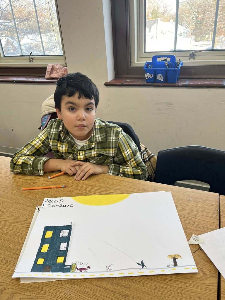 Young boy sitting at a desk with a drawing in front of him on the desk. 