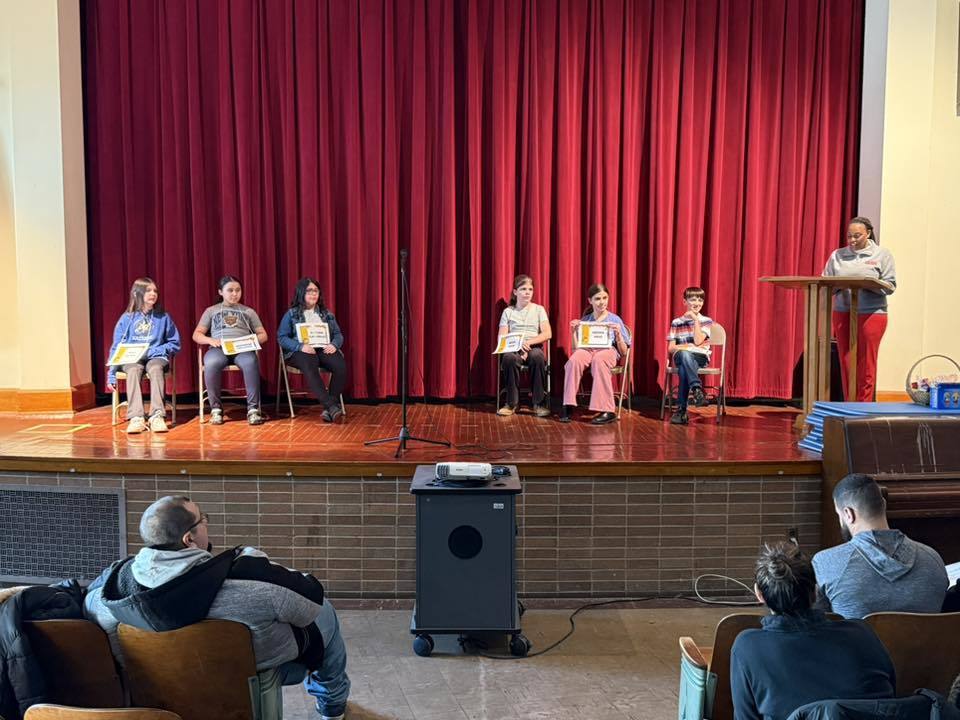 Kids on stage sitting against a red curtain and adult is standing at a podium.