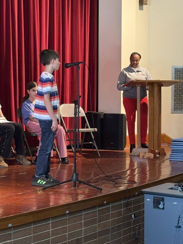 young boy standing at a microphone as an adult stands at a podium.