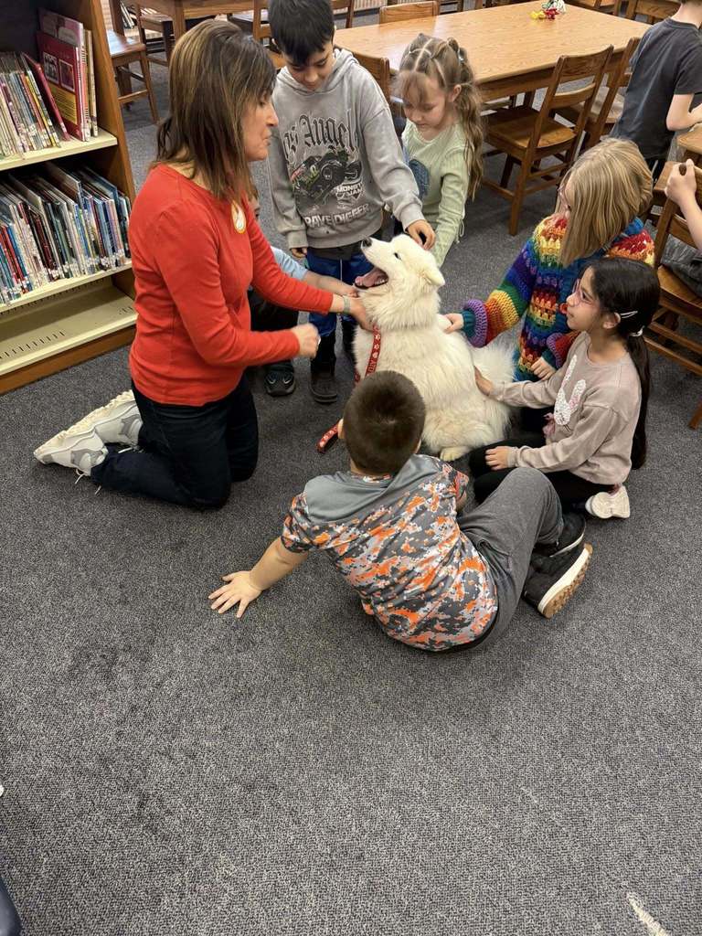 Women in a red shirt and students petting a white dog in a library.