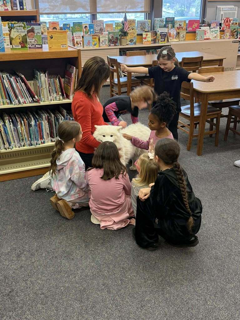 Women in a red shirt and students petting a white dog in a library.