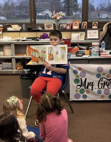 Young boy sitting at the front of a classroom and reading to students.