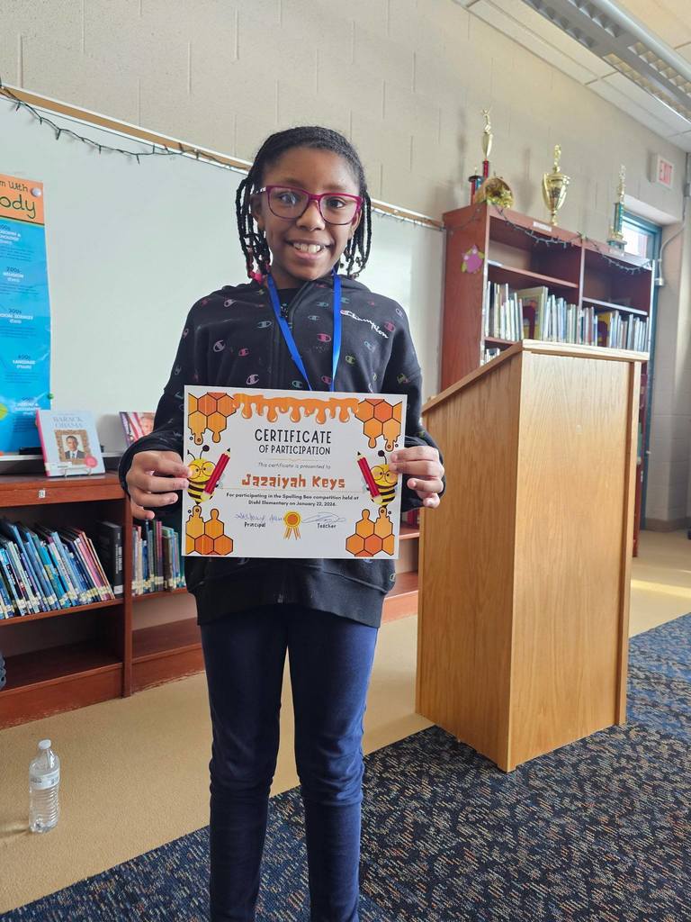 Young girl in a black patterned top and red glasses holding her certificate.