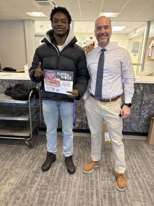 A Young man in a black coat with his pointer finger up standing next to a man with kaki pants and a tie. 