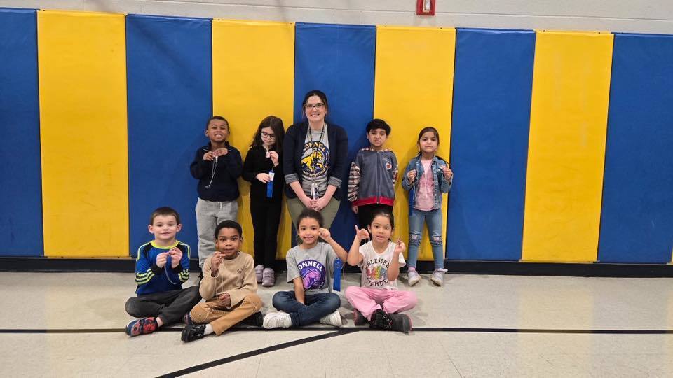 A group of kids with an adult , in front of a blue and yellow wall.