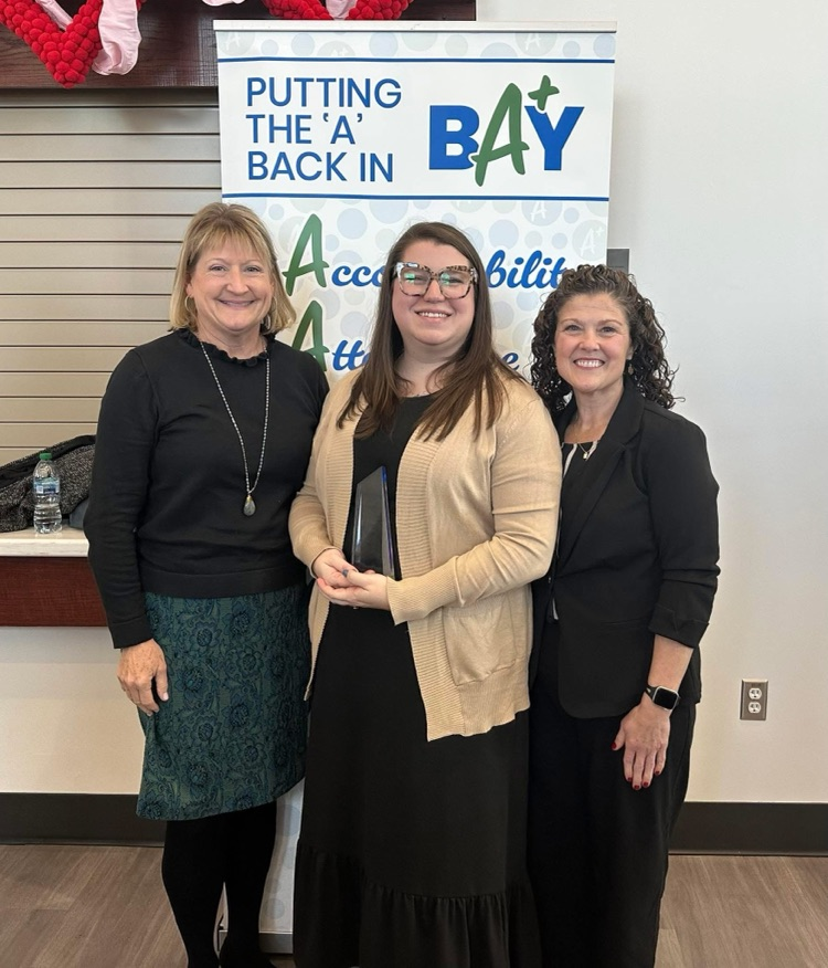 Three women smile while standing in front of a Bay District Schools banner; the woman in the center holds an award.