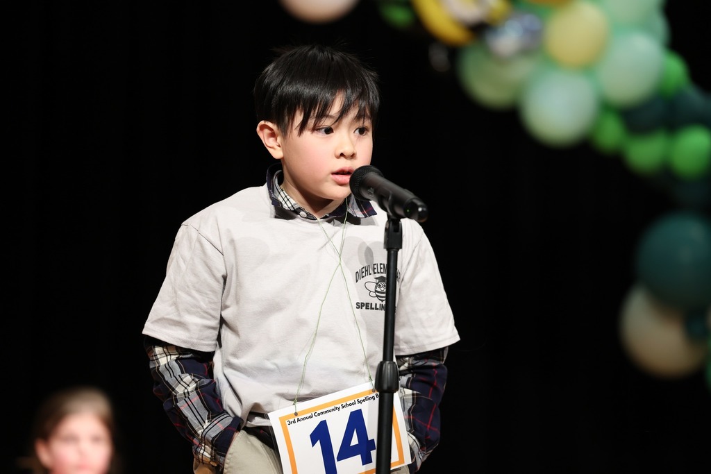 Young boy in a light gray shirt speaking into a microphone. 