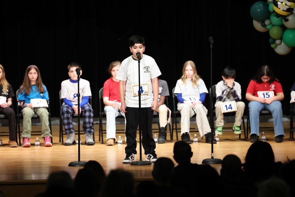 Young boy in a white shirt speaking into a microphone and standing on stage. 