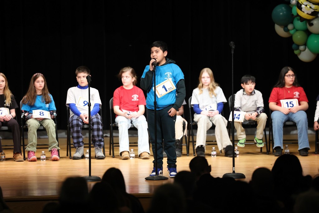 Young boy in a blue shirt speaking into a microphone and standing on stage. 
