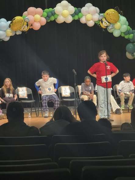 Young girl wearing a red shirt and speaking into a microphone on stage.