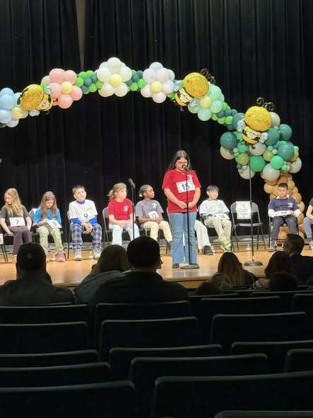 Young girl wearing a red shirt and speaking into a microphone on stage.