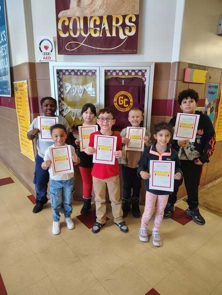A group of kids smiling in the hall and holding up white paper.