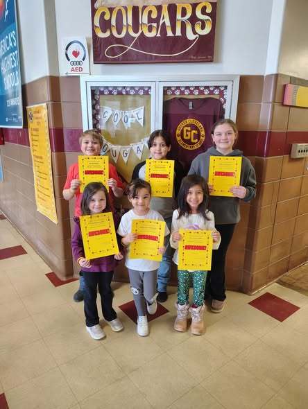 A group of kids smiling in the hall and holding up yellow paper.