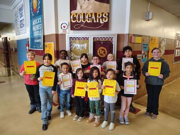 A group of kids smiling in the hall and holding up yellow and white paper.