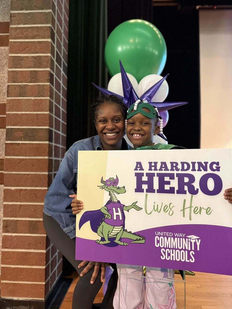 A young boy  standing next to a women in a jean jacket and smiling, while they hold a "Harding Hero" sign. 