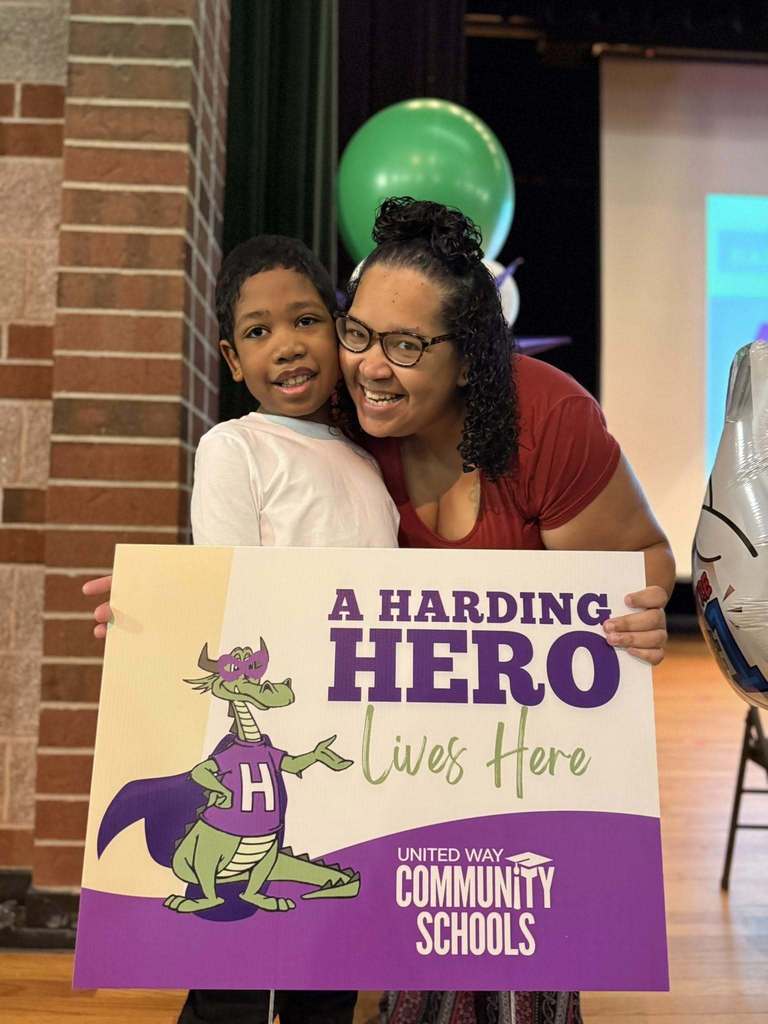A young boy in a white shirt standing next to a women in a red shirt and smiling, while they hold a "Harding Hero" sign. 