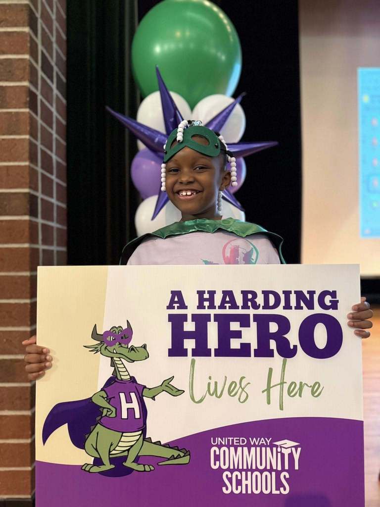 Young boy  wearing a green cape and holding a "Harding Hero" Sign. 