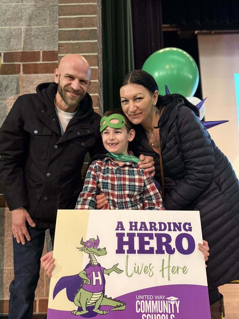 A man and women wearing a jacket standing next to a boy in a plaid shirt and is holding a "Harding Hero" Sign. 