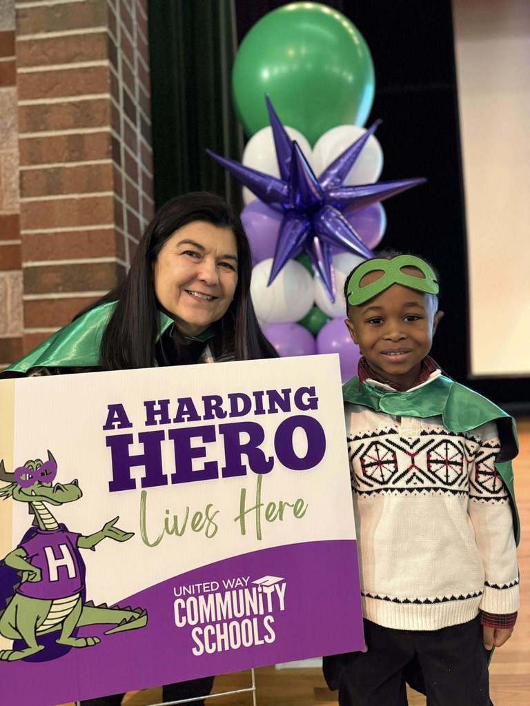 Women standing next to a young boy who is wearing a green cape with the "Harding Hero" sign in front of the lady. 