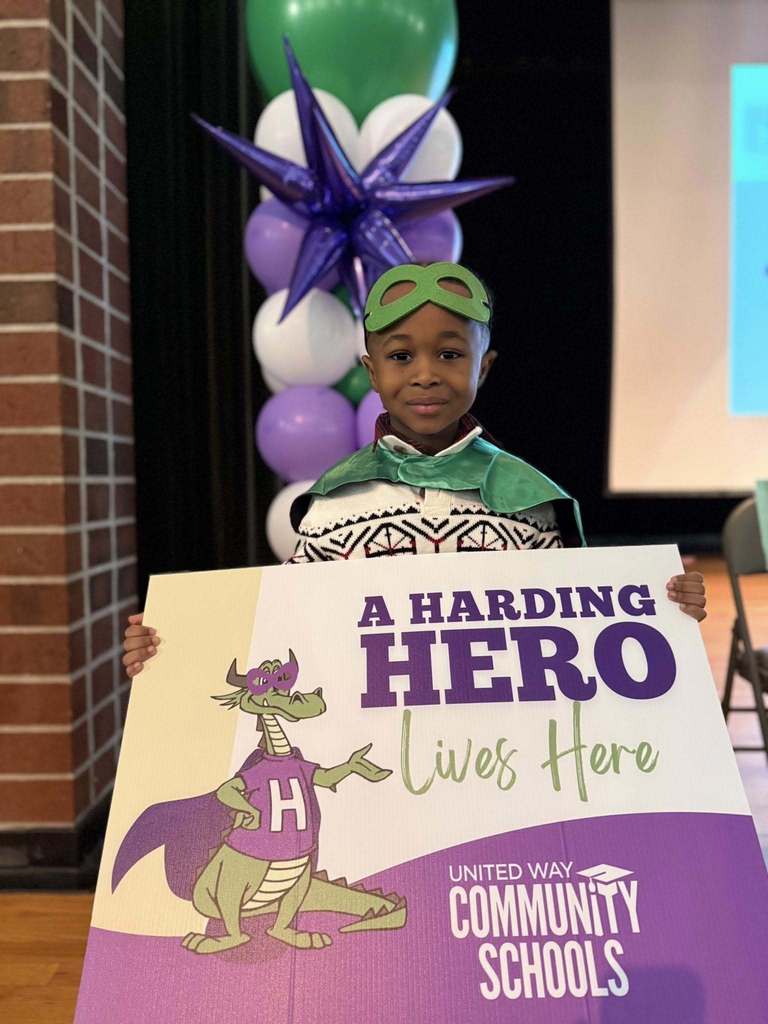 Young boy  wearing a green cape and holding a "Harding Hero" Sign. 