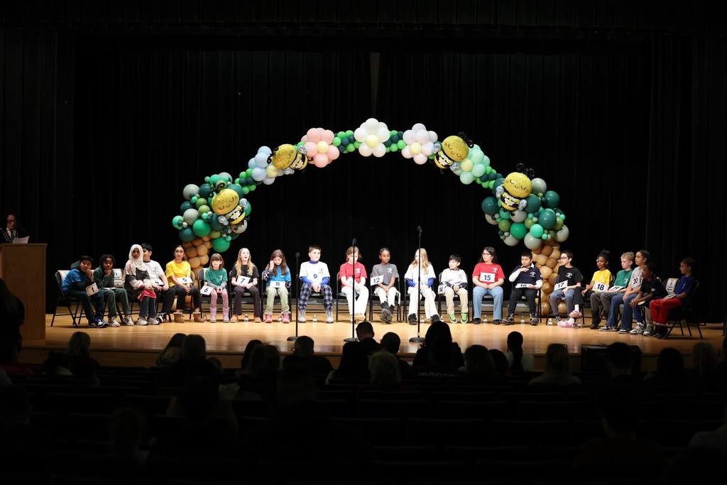 Kids sitting on a stage with a balloon arch behind them.