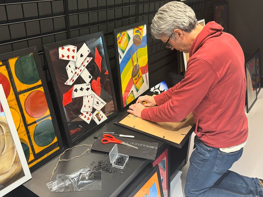 A man in a salmon colored hoodie putting art into picture frames. 