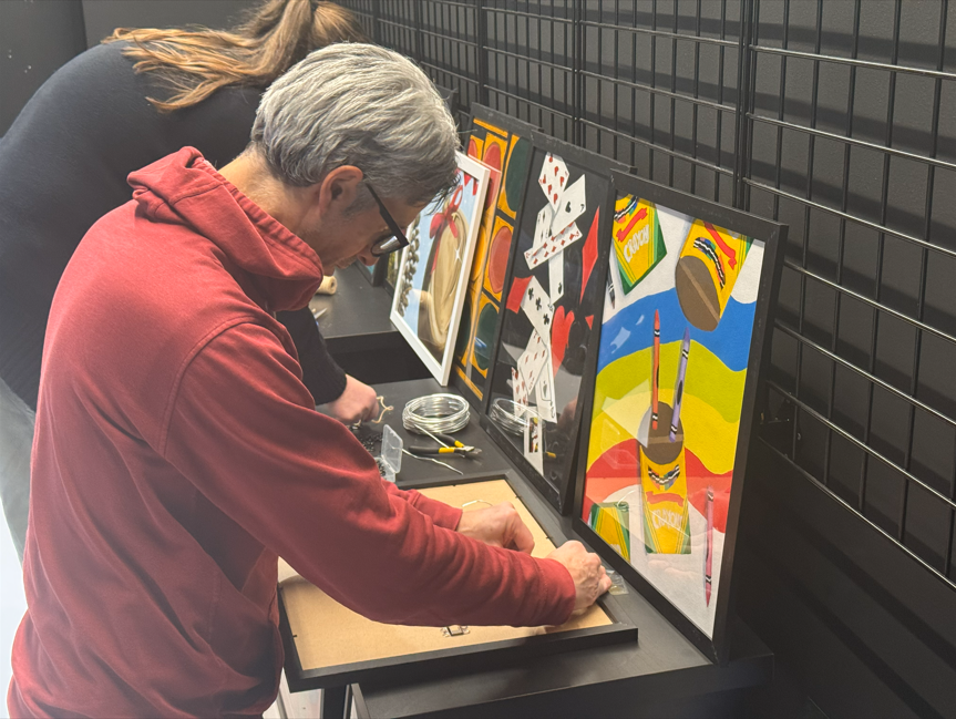 A man in a salmon colored hoodie putting art into picture frames. 