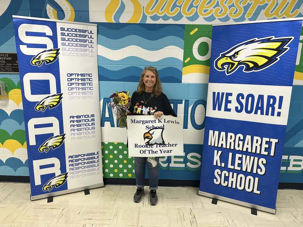 Teacher holding a ‘Margaret K. Lewis School Rookie Teacher of the Year’ sign and flowers, standing between school banners.