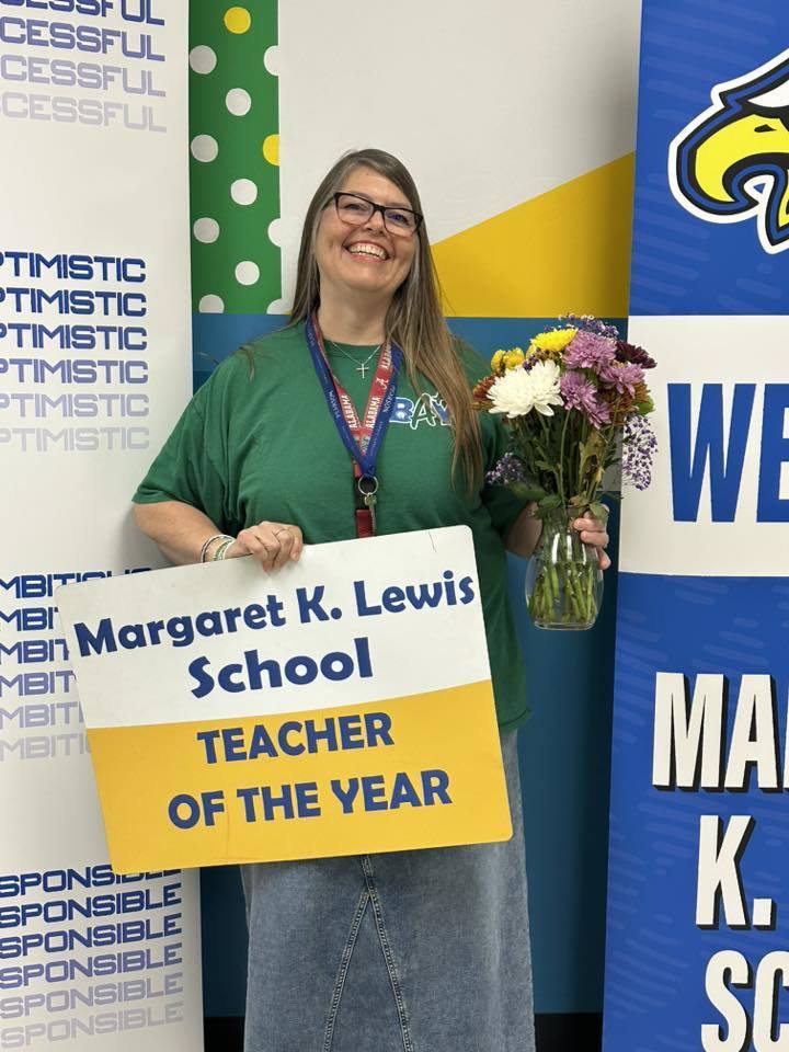 Smiling teacher holding a ‘Margaret K. Lewis School Teacher of the Year’ sign and a bouquet of flowers.