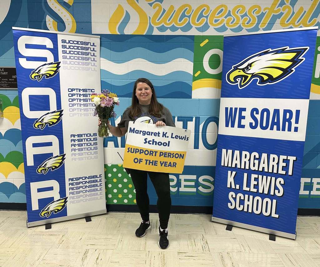 Staff member holding a ‘Margaret K. Lewis School Support Person of the Year’ sign and flowers, standing between school banners.