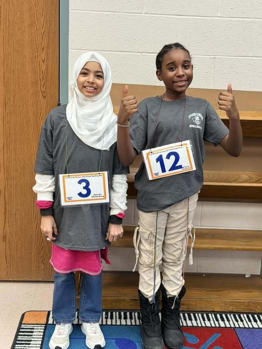 Two young girls smiling, in gray shirts and numbers hanging down from their shirts.  