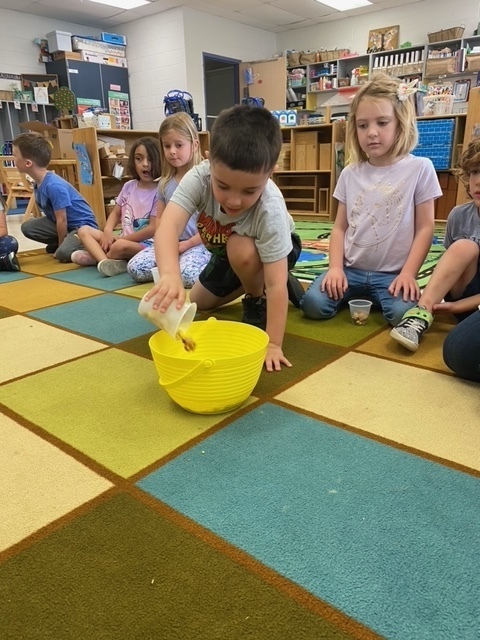 A little boy is adding a cup of food into a bowl to make a class trail mix.